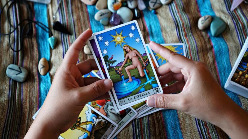 Mains tenant une carte de tarot représentant l’Étoile, posées sur un tissu coloré, entourées d’autres cartes de tarot et de pierres gravées de symboles, dans une ambiance introspective et créative.
