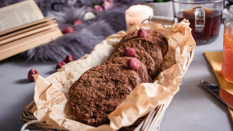 Biscuits au chocolat faits maison dans un panier, accompagnés d’un livre ouvert, d’une tasse de thé et d’une bougie, évoquant un moment de lecture réconfortant, de détente et de bien-être inspiré par la bibliothérapie.