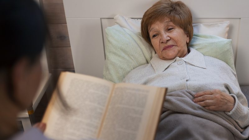 Une bibliothérapeute lit à voix haute un livre à une patiente âgée allongée dans son lit, dans un cadre de soin chaleureux.